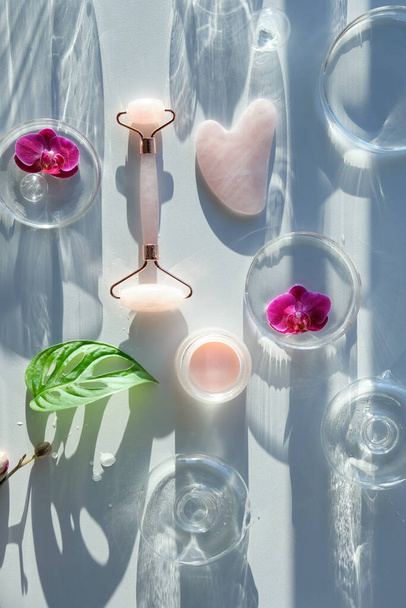Pink quartz face roller and guasha stone. Exotic monstera leaves, orchid flowers, petri dishes and glass balls. Sunlight, long shadows. Flat lay, off white background. Face lymph drainage background. - Фото, зображення