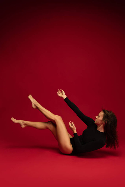 Young beautiful contemp female dancer, ballerina in art performance isolated on dark red background. Art, beauty, inspiration concept. - Foto, Imagen