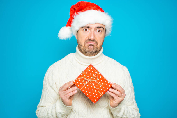 Προσωπογραφία ενός αηδιασμένου άνδρα που κρατάει κόκκινο κουτί δώρου σε μπλε φόντο φορώντας καπέλο Άη Βασίλη - Φωτογραφία, εικόνα