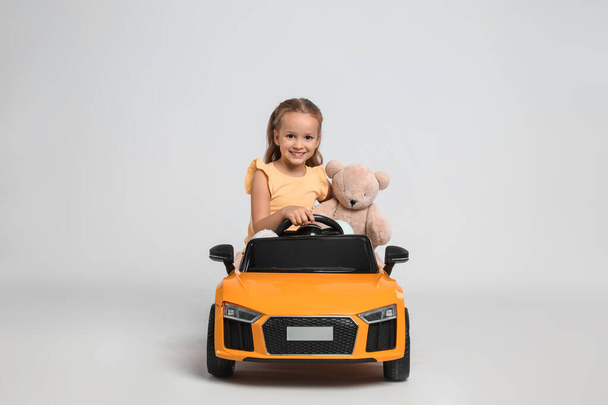 Cute little girl with toy bear driving children's car on grey background - Фото, изображение