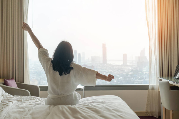 Χαλάρωση του ξενοδοχείου την τεμπέλικη μέρα με Ασιάτισσα γυναίκα ξυπνάει από τον καλό ύπνο στο κρεβάτι το πρωί του Σαββατοκύριακου αναπαύεται στο υπνοδωμάτιο άνεση κοιτάζοντας προς τη θέα της πόλης, έχοντας ευτυχισμένη, την ποιότητα ζωής της εργασίας ισορροπία τρόπο ζωής - Φωτογραφία, εικόνα