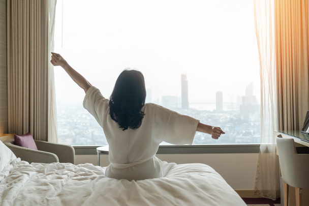 Hotel relaxation on lazy day with Asian woman waking up from good sleep on bed in weekend morning resting in comfort bedroom looking toward city view, having happy, work-life quality balance lifestyle - Foto, afbeelding