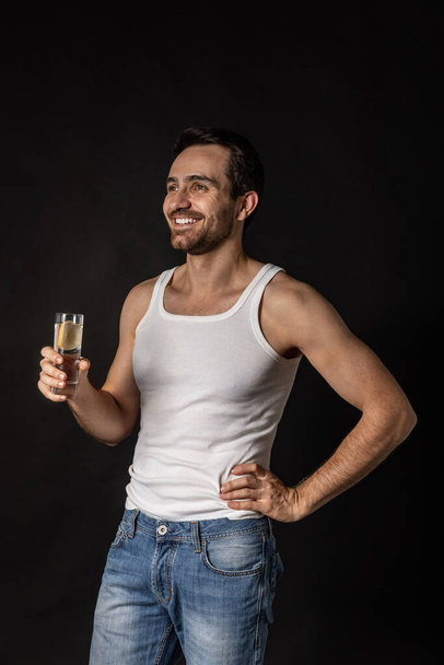 Classic portrait of young handsome bearded smiling man in white undershirt and jeans drinking water isolated on dark background. - Zdjęcie, obraz