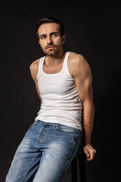 Classic portrait of young handsome bearded sad man in white undershirt and jeans isolated on dark background. Emotions, facial expression - Foto, Bild