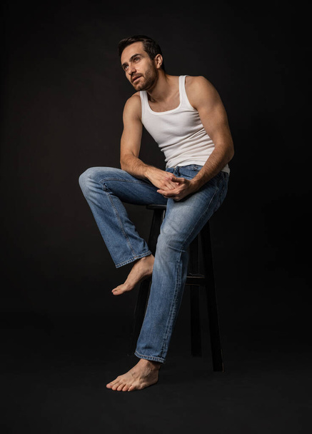 Classic portrait of young handsome bearded sad man in white undershirt and jeans isolated on dark background. Emotions, facial expression - Foto, Bild