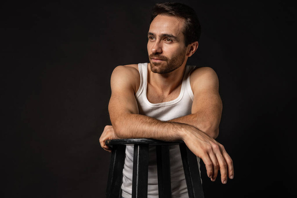 Classic portrait of young handsome bearded sad man in white undershirt and jeans isolated on dark background. Emotions, facial expression - Foto, Imagem