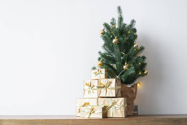 ein kleiner Weihnachtsbaum mit warmen Glühbirnen in einem Topf auf einem Holztisch mit Geschenken in Goldpapier gewickelt. das Konzept von Weihnachten. Hochwertiges Foto - Foto, Bild