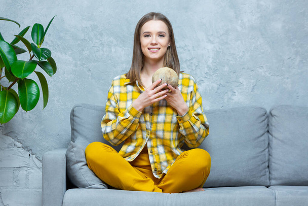 Front view of a smiling beautiful girl sitting cross legged on a sofa at home holding a coconut in both hands. Concept of healthy lifestyle, nutrition, detox, vegetarianism - Foto, Bild