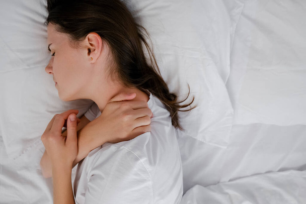 Top view of tired young brunette female lying on bed stretching and rubbing stiff shoulder muscles after waking up in morning. Upset woman feels discomfort because of bad mattress or posture problems - Φωτογραφία, εικόνα