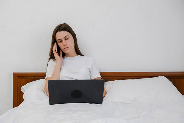 Young woman talking on phone using laptop sit on comfortable bed at home, caucasian female customer make mobile call confirming online website shopping order delivery concept, working remotely online - Foto, immagini