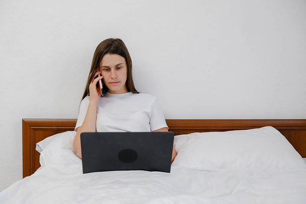 Young lady talking on smartphone using laptop sitting on comfy bed at home, cute female customer make mobile call confirming online website shopping order delivery concept, working remotely online - Valokuva, kuva