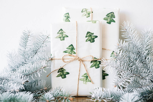 Новогодний подарок упакован в праздничную бумагу с акварельными елками на деревянном столе напротив белой стены с белыми еловыми ветвями. Высокое качество фото - Фото, изображение