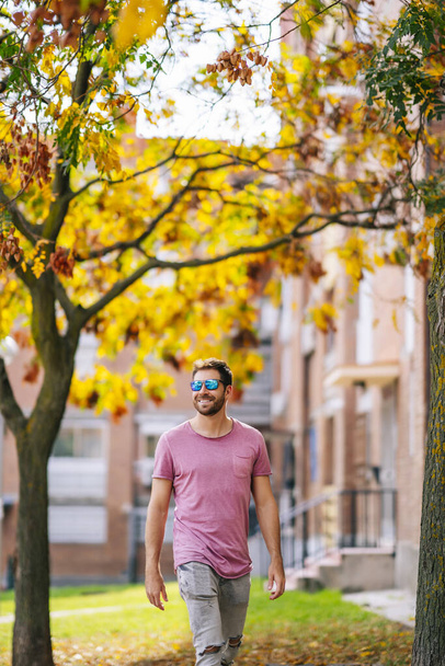 A young Caucasian stylish man wearing a nice pink t-shirt and sunglasses walking on a street - Foto, Bild