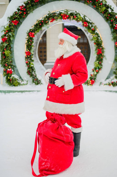 Santa Claus posing with a bag of gifts on the background of Christmas decorations outdoors - Фото, изображение