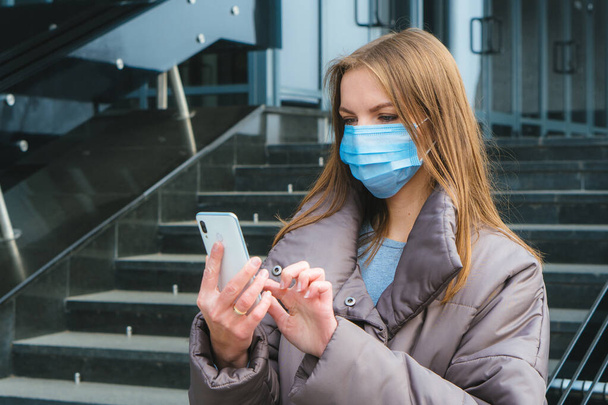 Joven mujer caucásica con máscara quirúrgica cara proteccionuse smartphone mientras camina en la calle exterior de la ciudad - Foto, Imagen