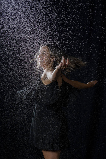 Μια σέξι γυναίκα με βρεγμένα μαλλιά φορώντας ένα μαύρο φόρεμα και απολαμβάνοντας το νερό πιτσιλιές σε ένα σκοτεινό στούντιο - Φωτογραφία, εικόνα