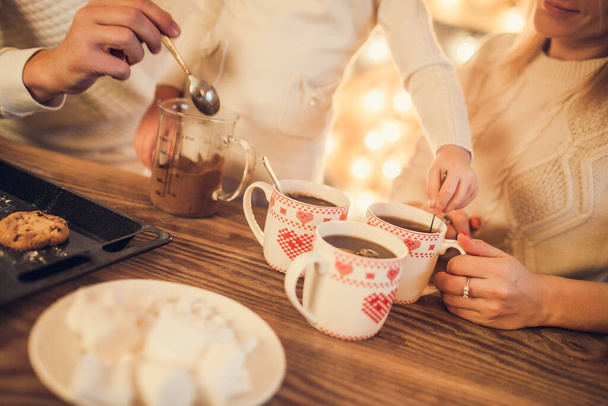 Famille : maman, papa et fille en pull blanc cuisinent et boivent du cacao avec des guimauves. Fermez les mains et les tasses. Concept de Noël. - Photo, image