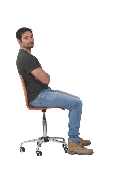 vista lateral de um retrato completo do homem sentado na cadeira olhando para câmera e braços cruzados no fundo whige - Foto, Imagem