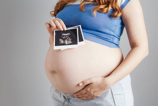smiling pregnant cute young caucasian woman with red hair and blue eyes standing against grey studio background and holding an ultrasound black and white scan, happy mother expectant - Foto, Imagen