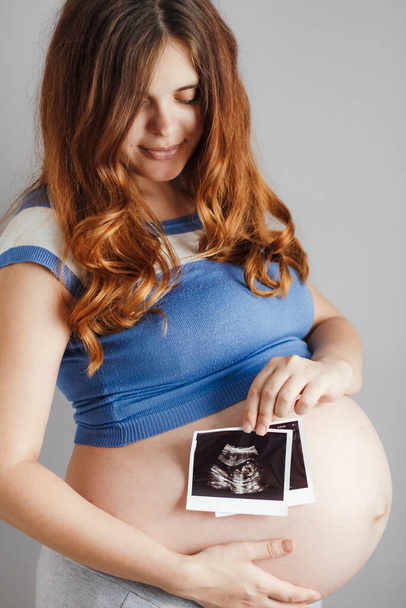 smiling pregnant cute young caucasian woman with red hair and blue eyes standing against grey studio background and holding an ultrasound black and white scan, happy mother expectant - Fotoğraf, Görsel