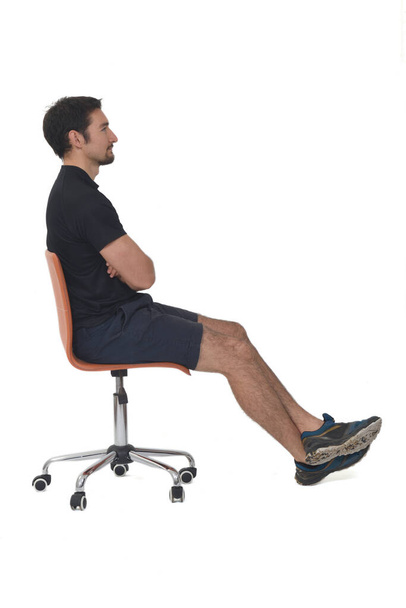 vista lateral de un retrato completo de un hombre sentado en una silla, usando ropa deportiva sobre fondo blanco - Foto, Imagen