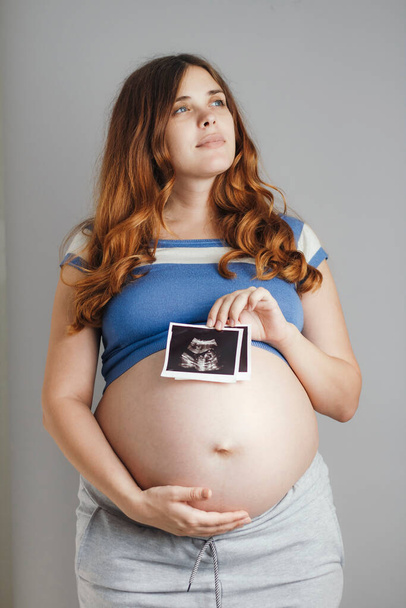beautiful pregnant young woman looking up on grey studio background and holding an ultrasound black and white scan, family life concept - Foto, Imagen