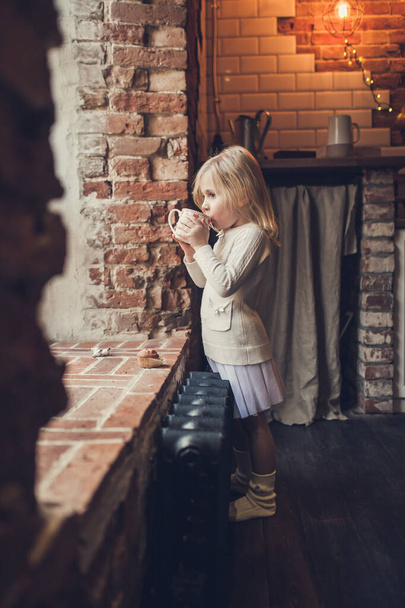 Ένα κοριτσάκι πίνει κακάο με ζαχαρωτά σε μια κουζίνα με έναν τοίχο από τούβλα και κοιτάζει έξω από ένα μεγάλο παράθυρο, σκέφτεται και ονειρεύεται. - Φωτογραφία, εικόνα