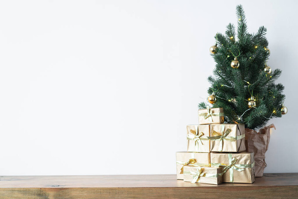 ein kleiner Weihnachtsbaum mit warmen Glühbirnen in einem Topf auf einem Holztisch mit Geschenken in Goldpapier gewickelt. das Konzept von Weihnachten. Hochwertiges Foto - Foto, Bild