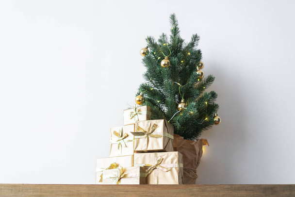 ein kleiner Weihnachtsbaum mit warmen Glühbirnen in einem Topf auf einem Holztisch mit Geschenken in Goldpapier gewickelt. das Konzept von Weihnachten - Foto, Bild