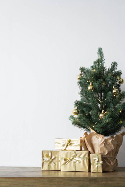 mała choinka z ciepłymi cebulkami w garnku na drewnianym stole z prezentami zawiniętymi w złoty papier. koncepcja Bożego Narodzenia. Wysokiej jakości zdjęcie - Zdjęcie, obraz