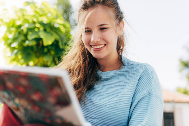 Uma estudante positiva lê um livro enquanto se senta no banco em um dia ensolarado. Mulher sorridente em roupa casual descansa do lado de fora, fazendo lição de casa e aprendendo com um livro. - Foto, Imagem