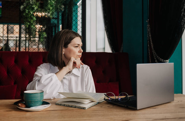Eine nachdenkliche brünette Geschäftsfrau sitzt mit Laptop und Notizbuch an einem Tisch in einem Café - Foto, Bild