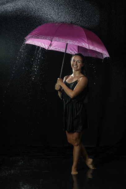 A dancer wearing a black dress, holding an umbrella, and enjoying the water splashes in a dark studio - Fotoğraf, Görsel
