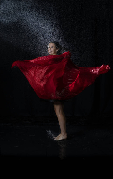 A dancer with wet hair wearing a black dress, enjoying the water splashes and dancing with a red scarf - Photo, image