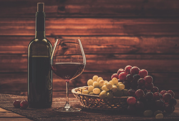 Bottle of red wine, glass and grape in basket in wooden interior  - Photo, image
