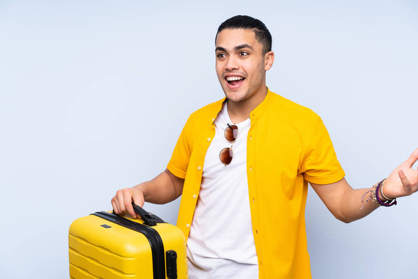 Asyalı yakışıklı adam mavi arka planda izole edilmiş seyahat valiziyle ve şaşırmış - Fotoğraf, Görsel