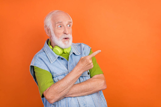 Photo of impressed shocked retired man wear jeans waistcoat headphones pointing looking empty space isolated orange color background - Foto, immagini