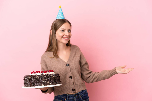 Mladá Litevka drží narozeninový dort izolované na růžovém pozadí natahuje ruce na stranu pro pozvání přijít - Fotografie, Obrázek