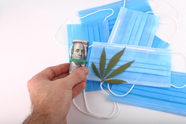 Folha de cannabis na mão contra o fundo de uma máscara médica de coronavírus e cem dólares americanos - Foto, Imagem