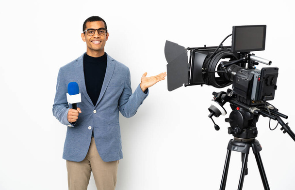 Репортёр афроамериканец держит микрофон и сообщает новости на изолированном белом фоне, держа на ладони воображаемое копирайт-пространство, чтобы поместить объявление - Фото, изображение