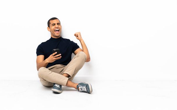 Hombre afroamericano sentado en el suelo con el teléfono en posición de victoria - Foto, imagen
