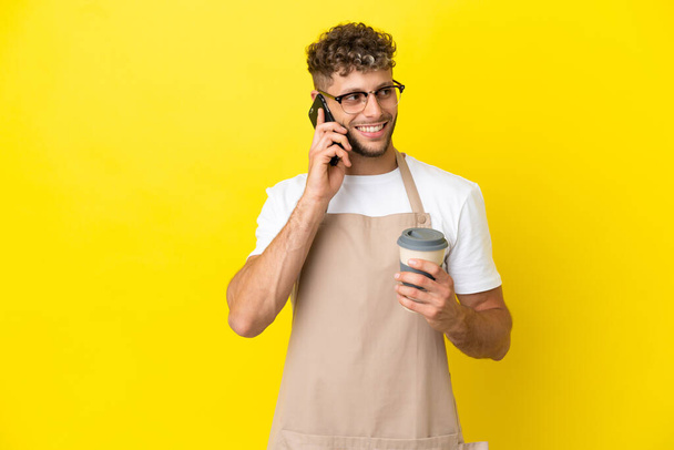 Étterem pincér szőke férfi elszigetelt sárga háttér gazdaság kávé elvitelre és egy mobil - Fotó, kép