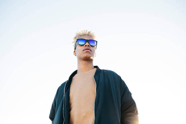 Joven hombre latinoamericano con gafas de sol azules contra un cielo blanco - Foto, Imagen