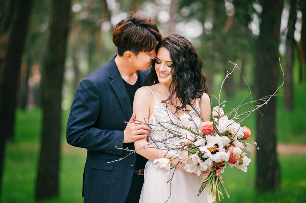 Την ημέρα του γάμου. Νέοι ευτυχισμένοι νιόπαντροι σε φρέσκο πράσινο δάσος. Διεθνές ζευγάρι. Πορτρέτο νύφης και γαμπρού. Ανοιξιάτικος γάμος. Εξωτερική γαμήλια τελετή. - Φωτογραφία, εικόνα