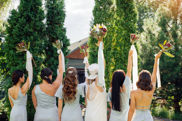 Bridesmaids in blue dresses and a bride in white gown with a hat holding beautiful bouquets of fresh protea flowers. Gorgeous luxury summer wedding, outdoor with green trees background, garden party. - Foto, imagen