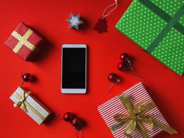 Joulun ja uudenvuoden lahjoja, gadget näyttö ja loma koristeet punaisella taustalla. Juhlakortti punaisella taustalla, joulukuusi koristeet, kynttilät, loma koristeet postikortti, älypuhelin näyttö, tabletti - Valokuva, kuva