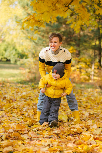 Happy mother and little son having fun together in autumn park among the yellow leaves - Фото, изображение