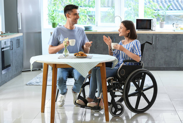 Jonge vrouw in rolstoel en haar man drinken thee in de keuken - Foto, afbeelding