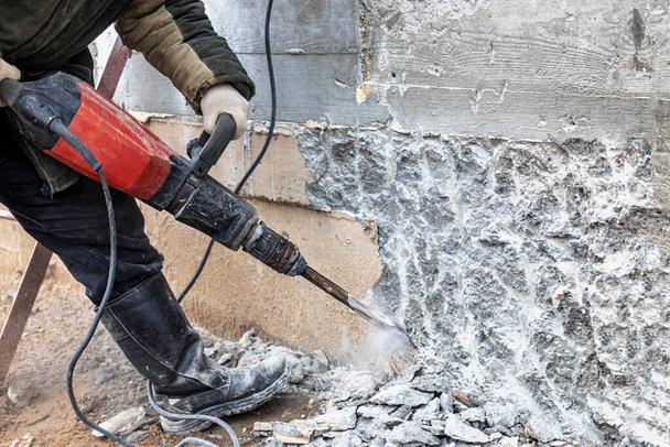 Un ouvrier avec un marteau-piqueur sur un chantier de construction nivelle un mur de béton. Puissant équipement industriel manuel. Travaux de construction difficiles - Photo, image