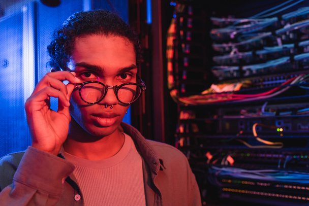 young african american engineer touching eyeglasses while looking at camera near blurred server - Photo, Image